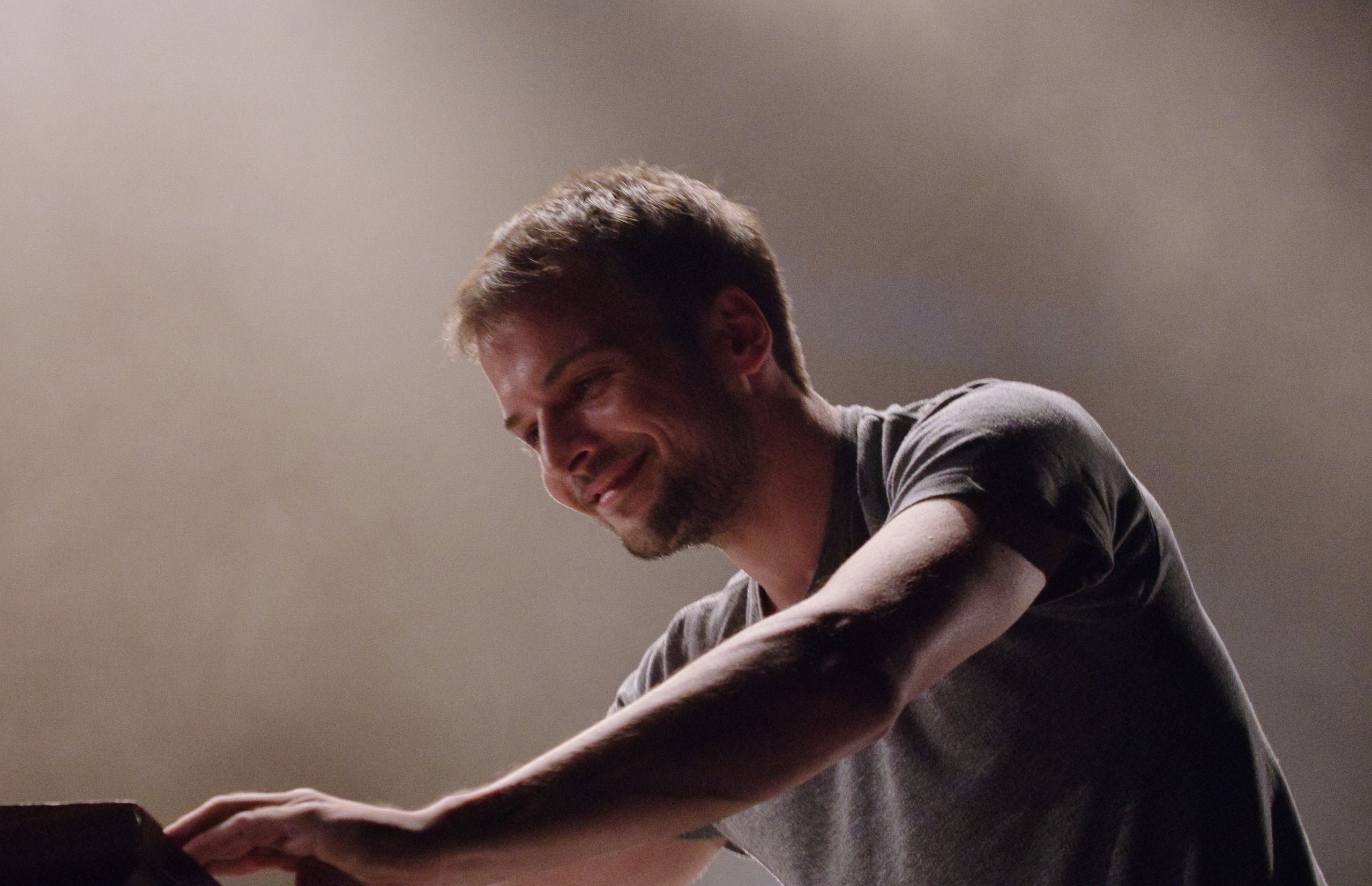 Young man with slight beard smiling while playing piano on stage.
