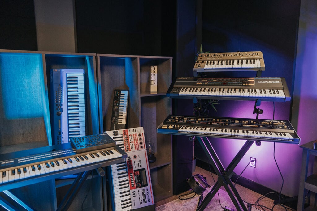 Synthesizer keyboards on racks and shelves in blue and purple lit studio.